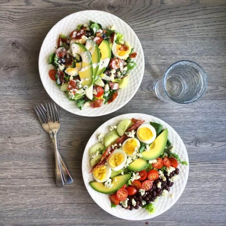 two cobb salads on a wooden table, glass of water on the right and fork on the left